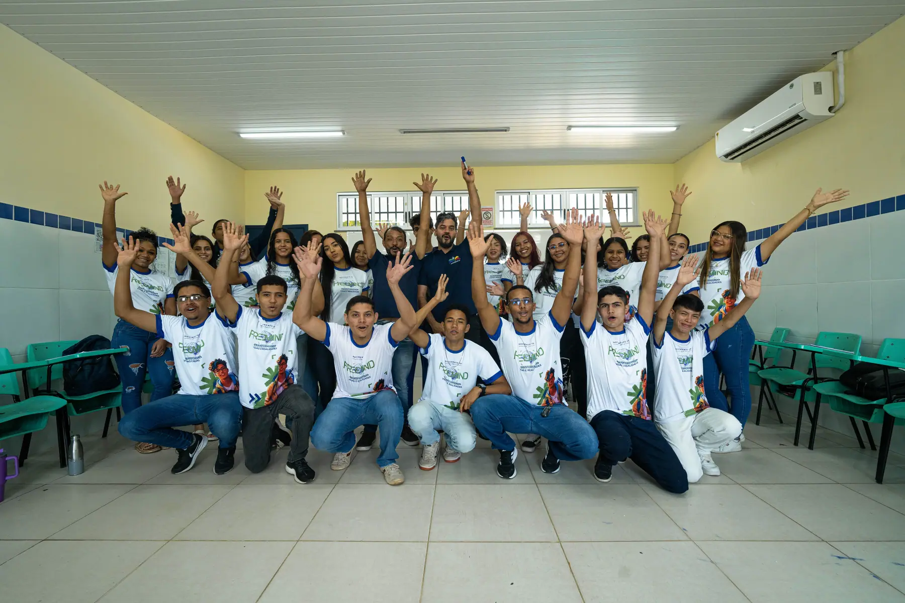Turma de estudantes e educadores do Movimento ID, de camisa branca, com os braços erguidos em uma sala de aula